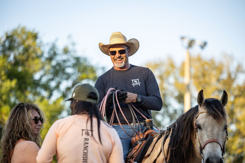 Smiling cowboy in hat and sunglasses rides horse at outdoor rodeo event with spectators nearby.