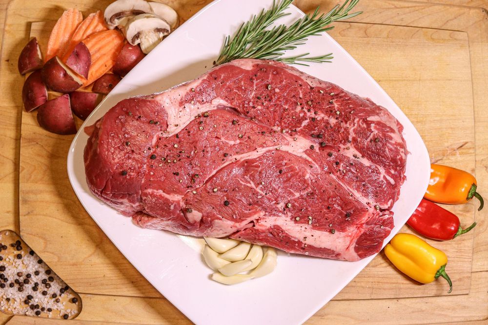 Raw ribeye steak seasoned with pepper on white plate, surrounded by colorful mini peppers, garlic, mushrooms, and rosemary.