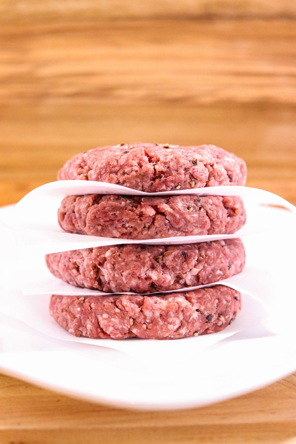 Stack of four raw ground beef burger patties separated by parchment paper on a white plate.