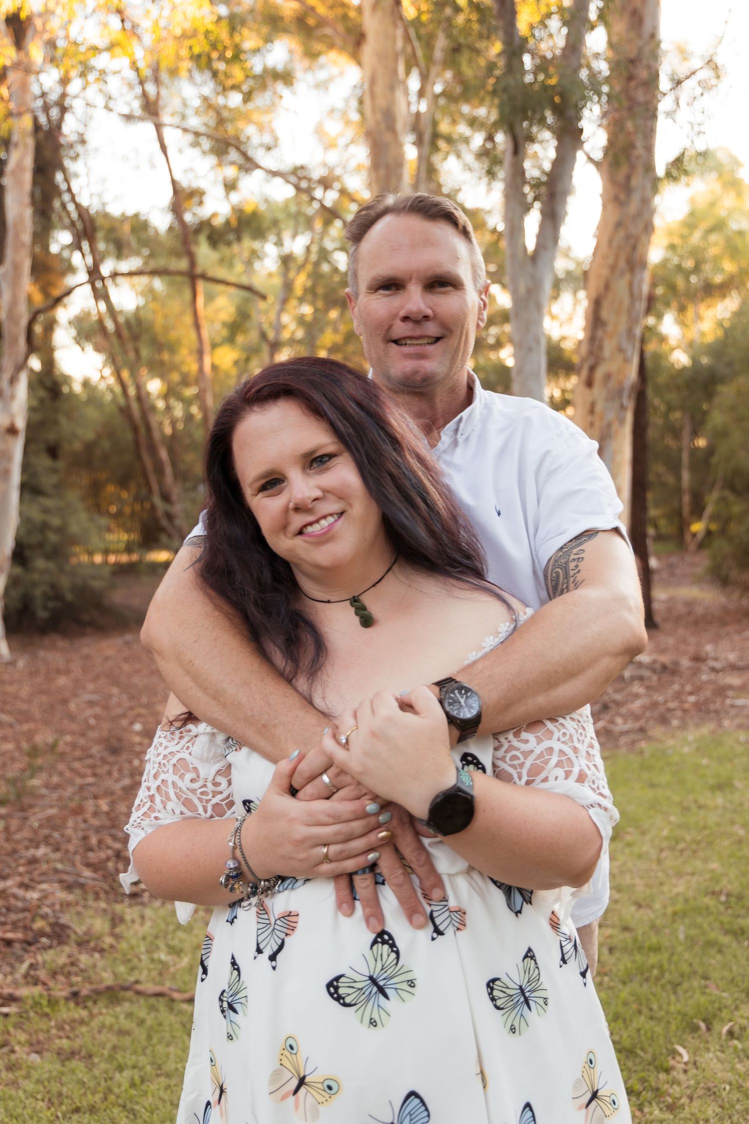 A smiling couple embraces outdoors in a park, woman in butterfly dress, man in white shirt, surrounded by trees.