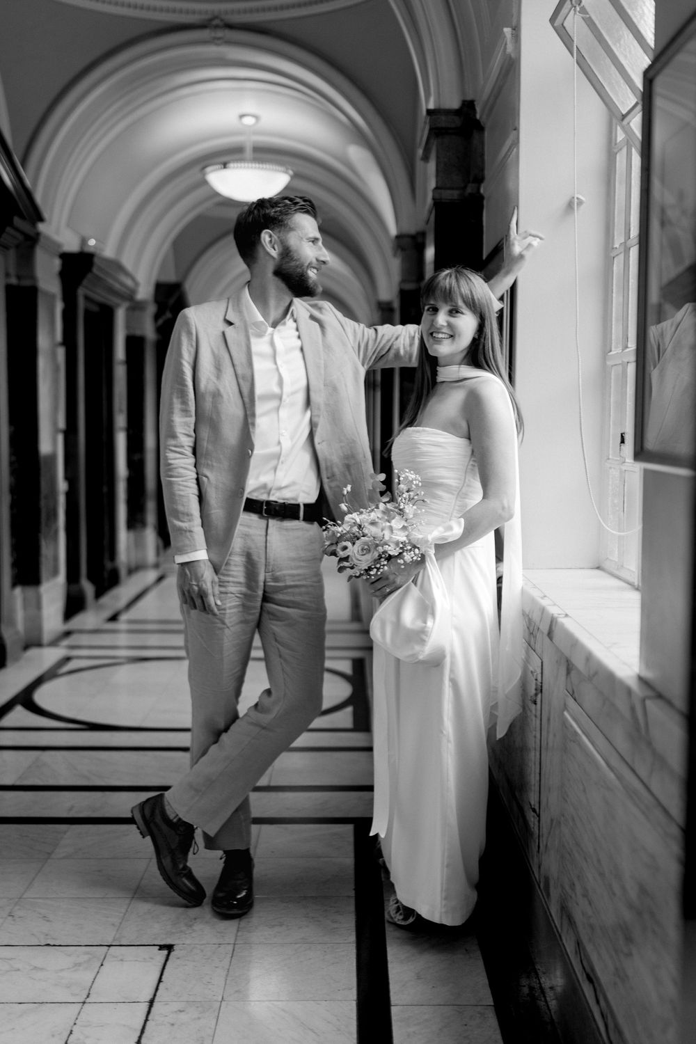 Black and white photo of a bride and groom walking through an elegant arched hallway, bride holding a bouquet.