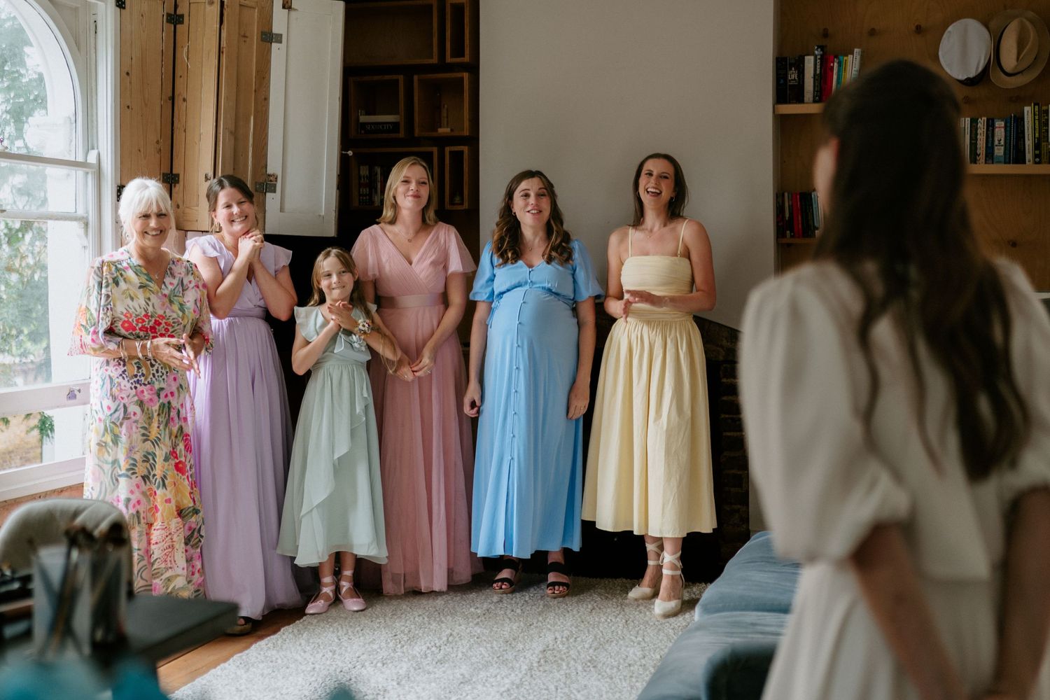Bridesmaids in pastel dresses stand together smiling as bride in white gown reveals her wedding look indoors.