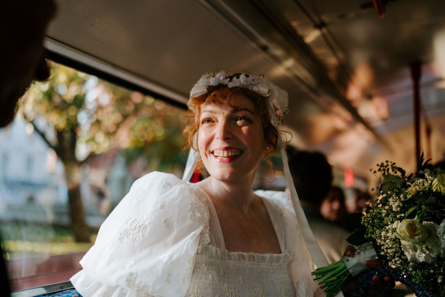 Smiling bride in white dress and floral headpiece holding bouquet while riding a trolley on her wedding day.