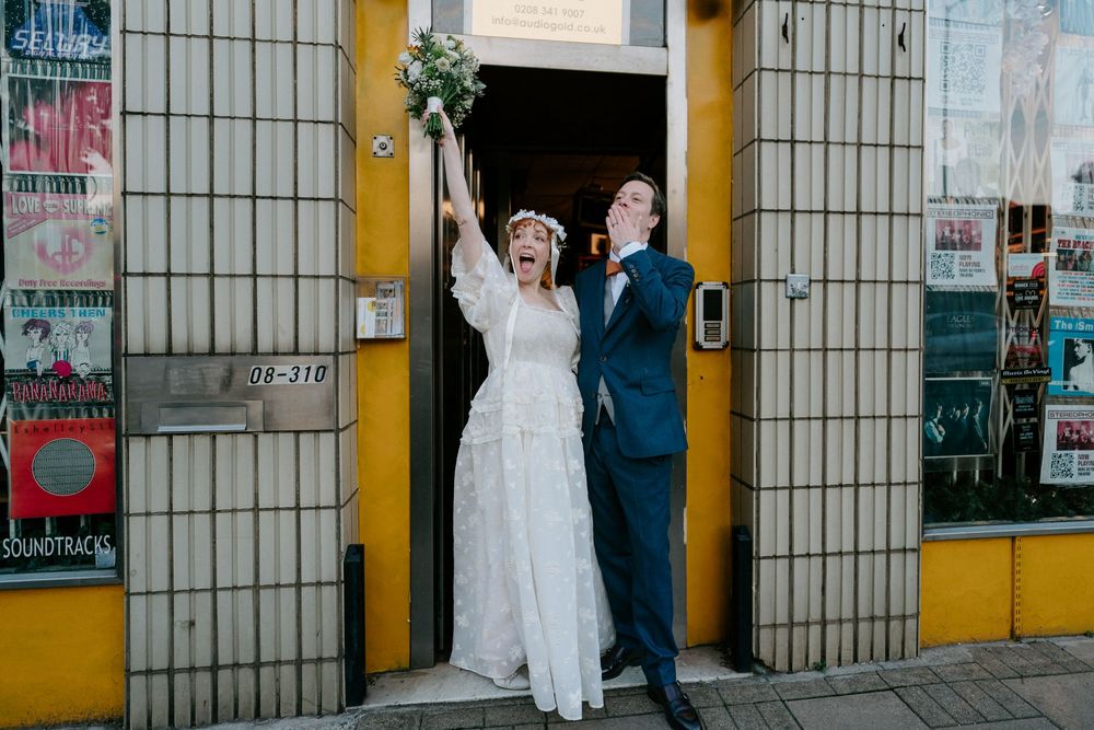 Joyful bride and groom celebrating outside a venue, bride raising bouquet, groom in teal suit, urban street backdrop.