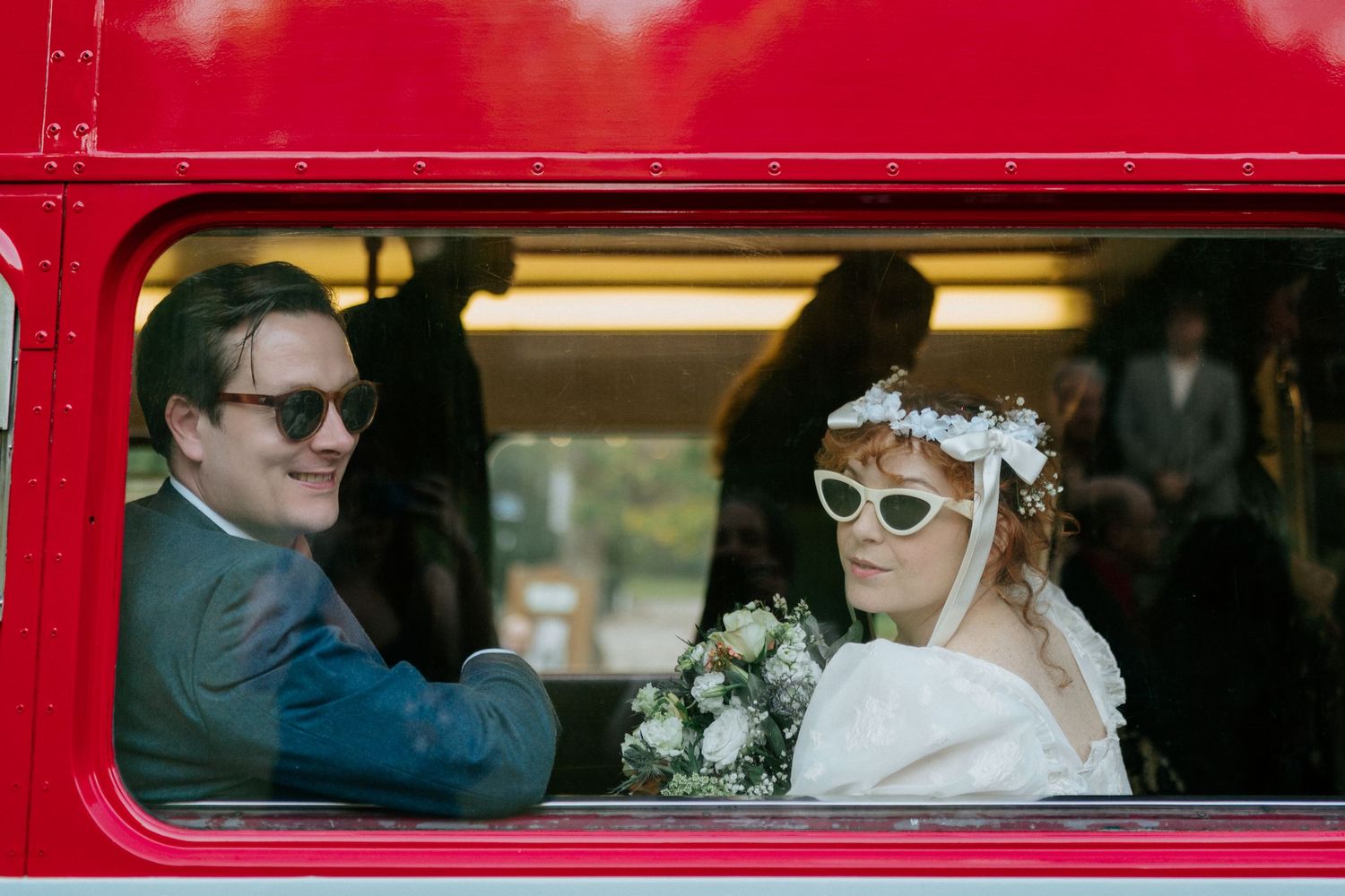 Bride and groom riding a red double-decker bus, wearing sunglasses, bride holding white floral bouquet with flower crown.