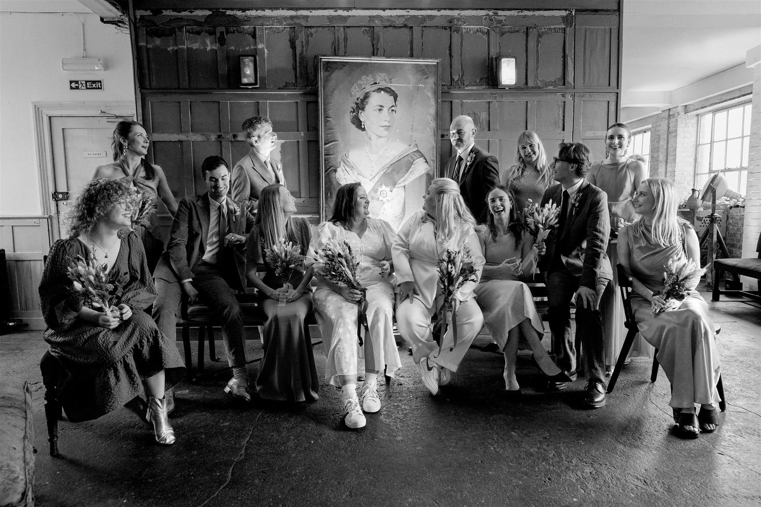 Black and white wedding party photo with bride, groom, and guests posed before a large portrait painting indoors.