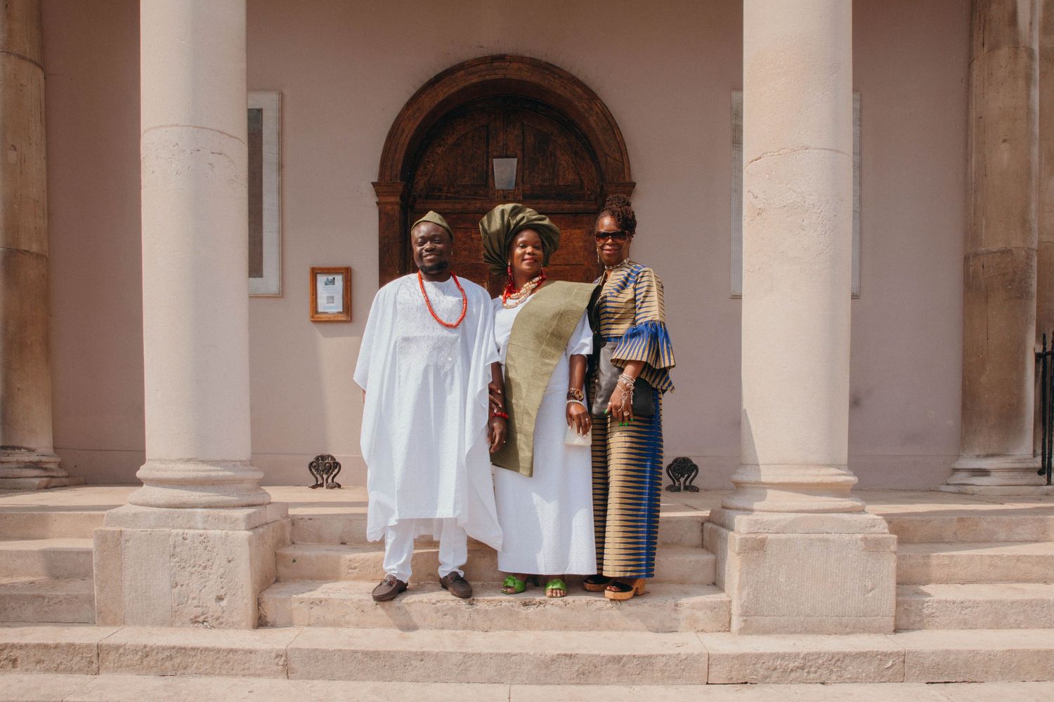 Three people in traditional African attire pose on steps before a historic building with columns and wooden door.
