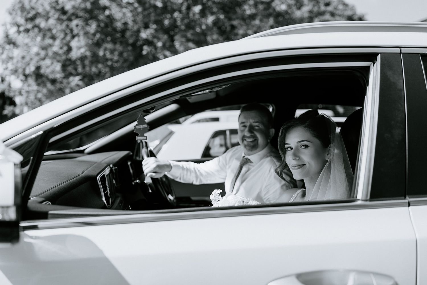 Black and white photo of a smiling couple in a white car, dressed in wedding attire, bride and groom on their special day.