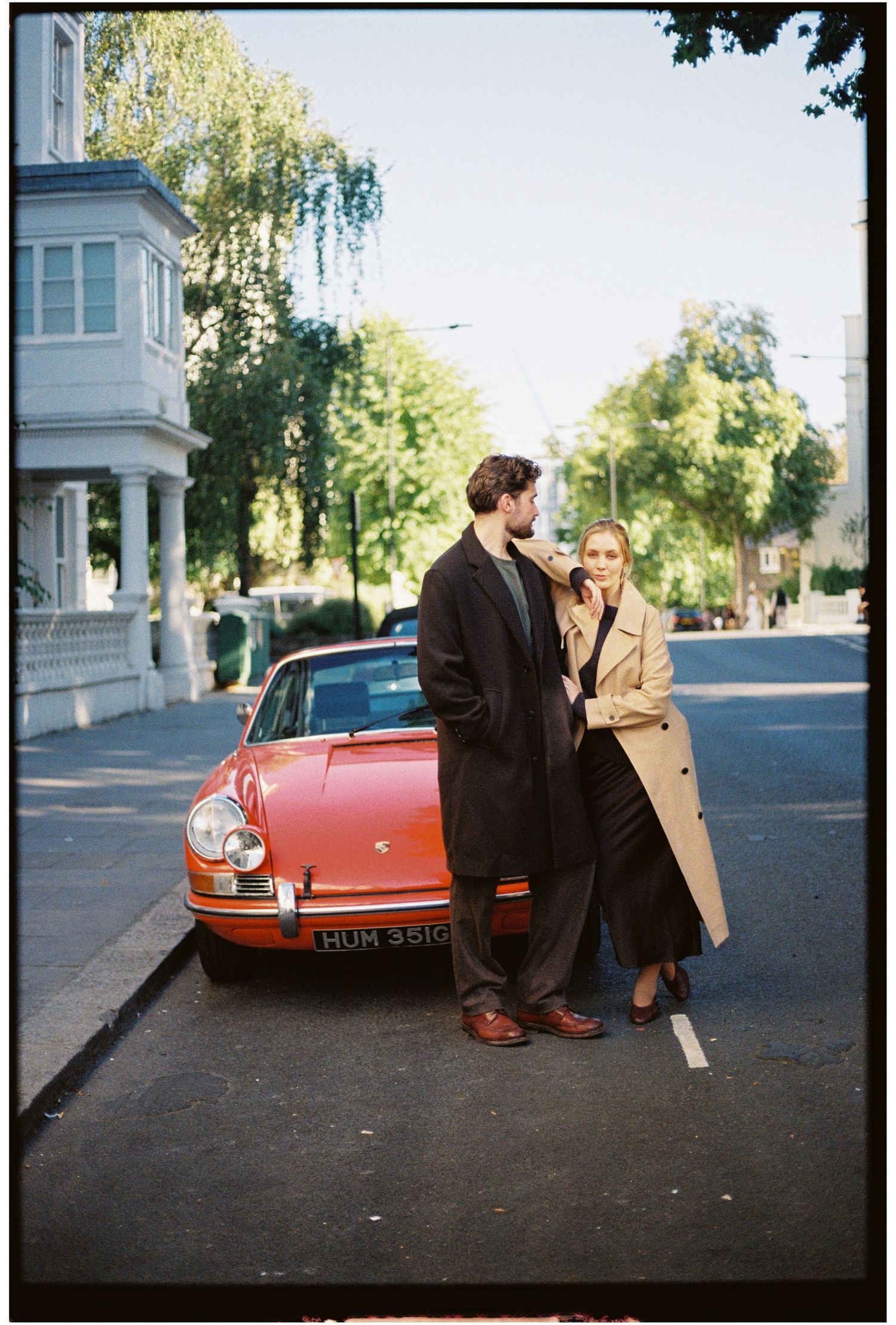 Couple in elegant coats leaning against a vintage red Porsche on a tree-lined street.