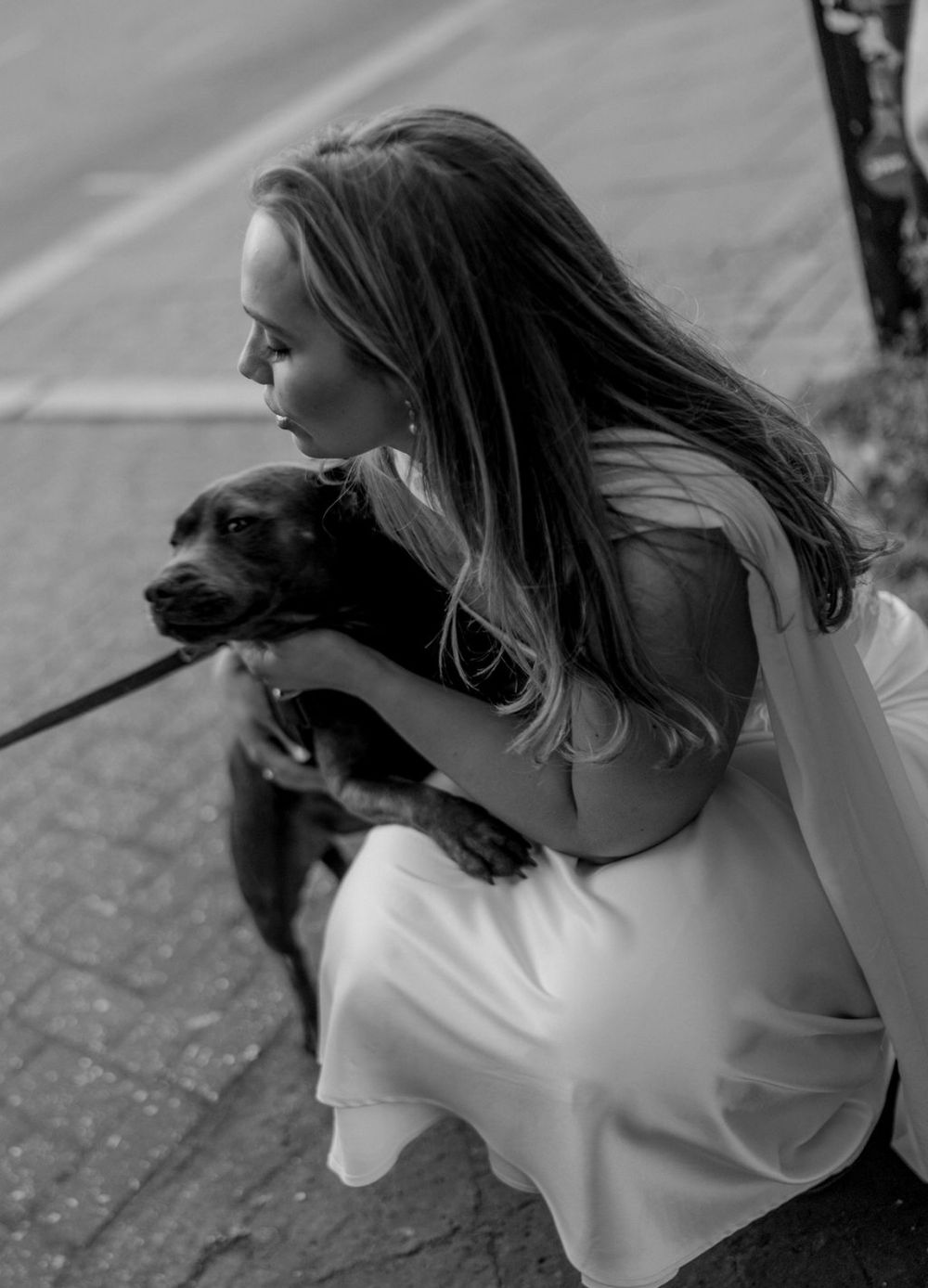 Bride in white dress crouching to embrace a small black dog on a city sidewalk, black and white photo.