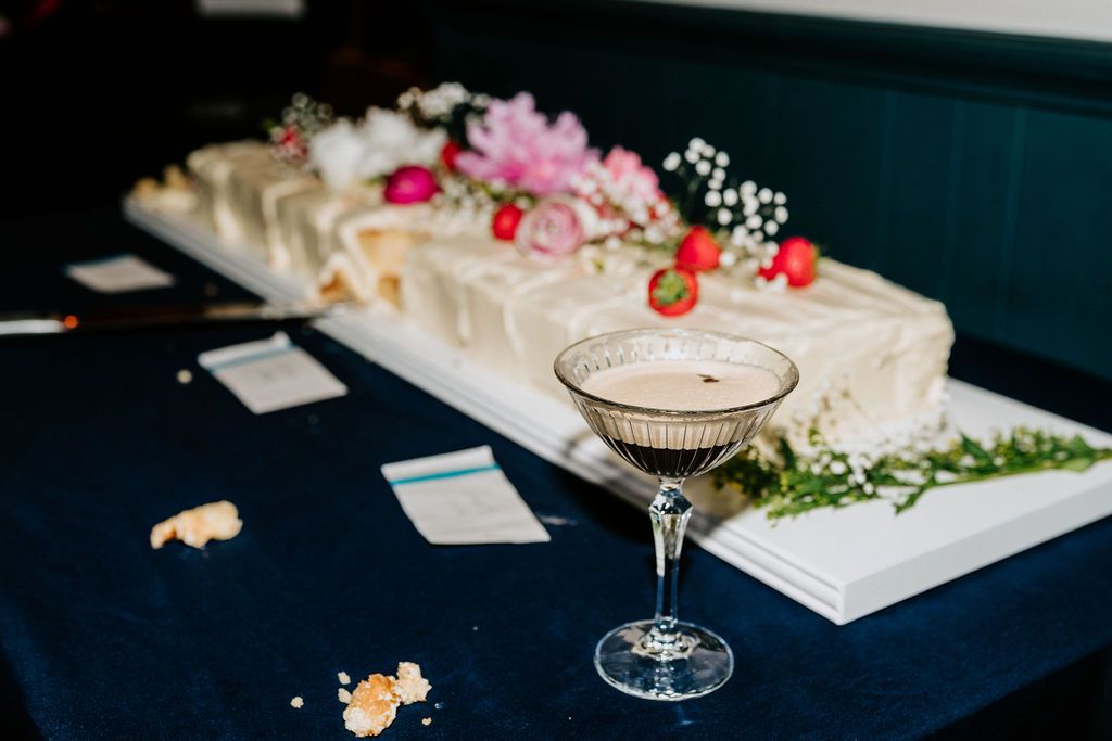 Elegant cocktail in coupe glass beside a decorated cake on a navy blue table with floral garnishes.