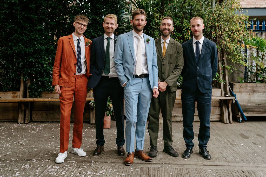 Five men in colorful suits pose outdoors, wearing shades of orange, navy, light blue, and tweed for a stylish group photo.