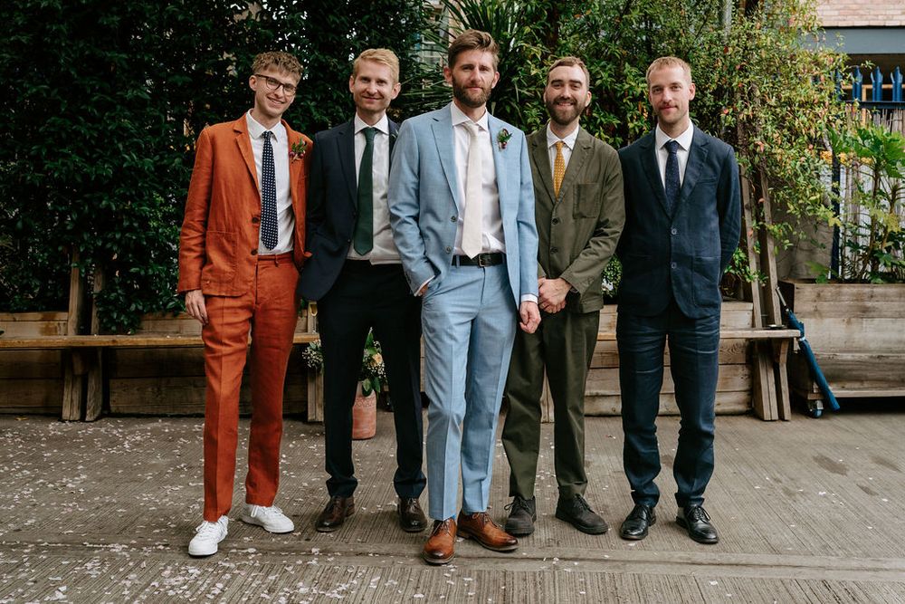 Five men in colorful suits pose outdoors, wearing shades of orange, navy, light blue, and tweed for a stylish group photo.