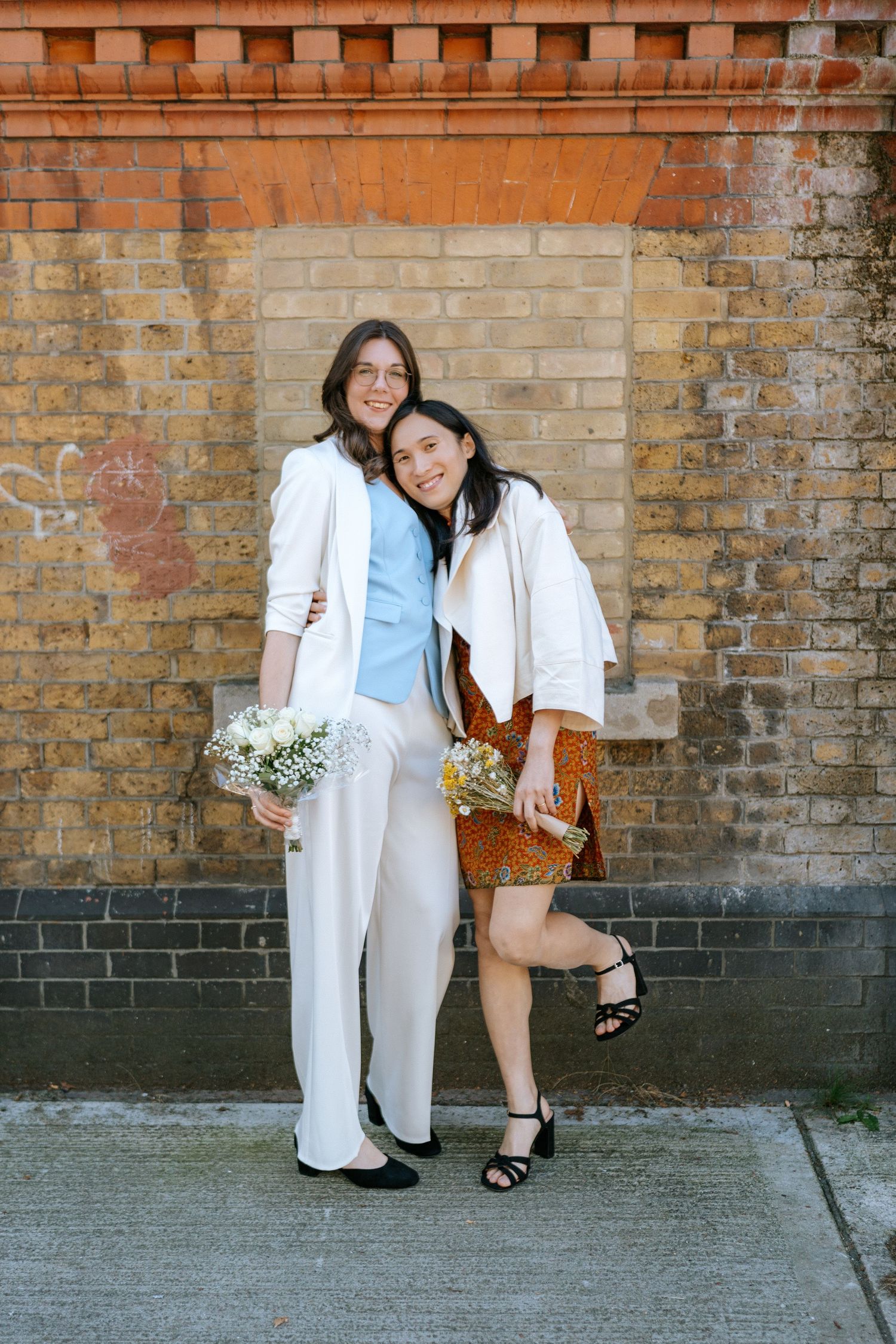 Two women pose smiling against a brick wall, one in white suit holding flowers, other in brown floral dress with small bouquet.