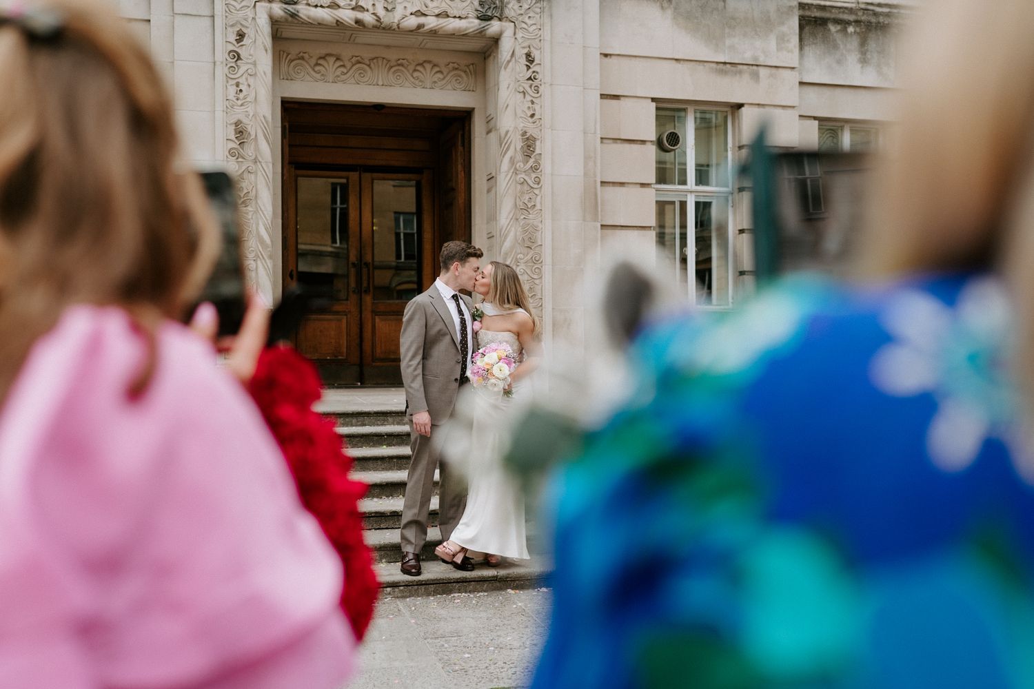 Couple sharing a kiss outside elegant building entrance while wedding guests in colorful attire watch in foreground.