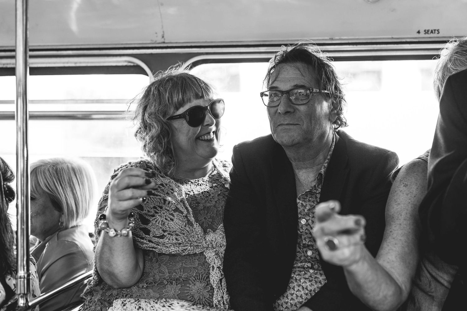 Black and white photo of a smiling couple sitting together on a bus, woman wearing sunglasses and lace top.