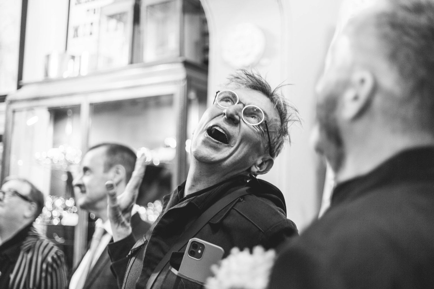 Black and white photo of a laughing woman wearing glasses holding a phone at a social gathering.