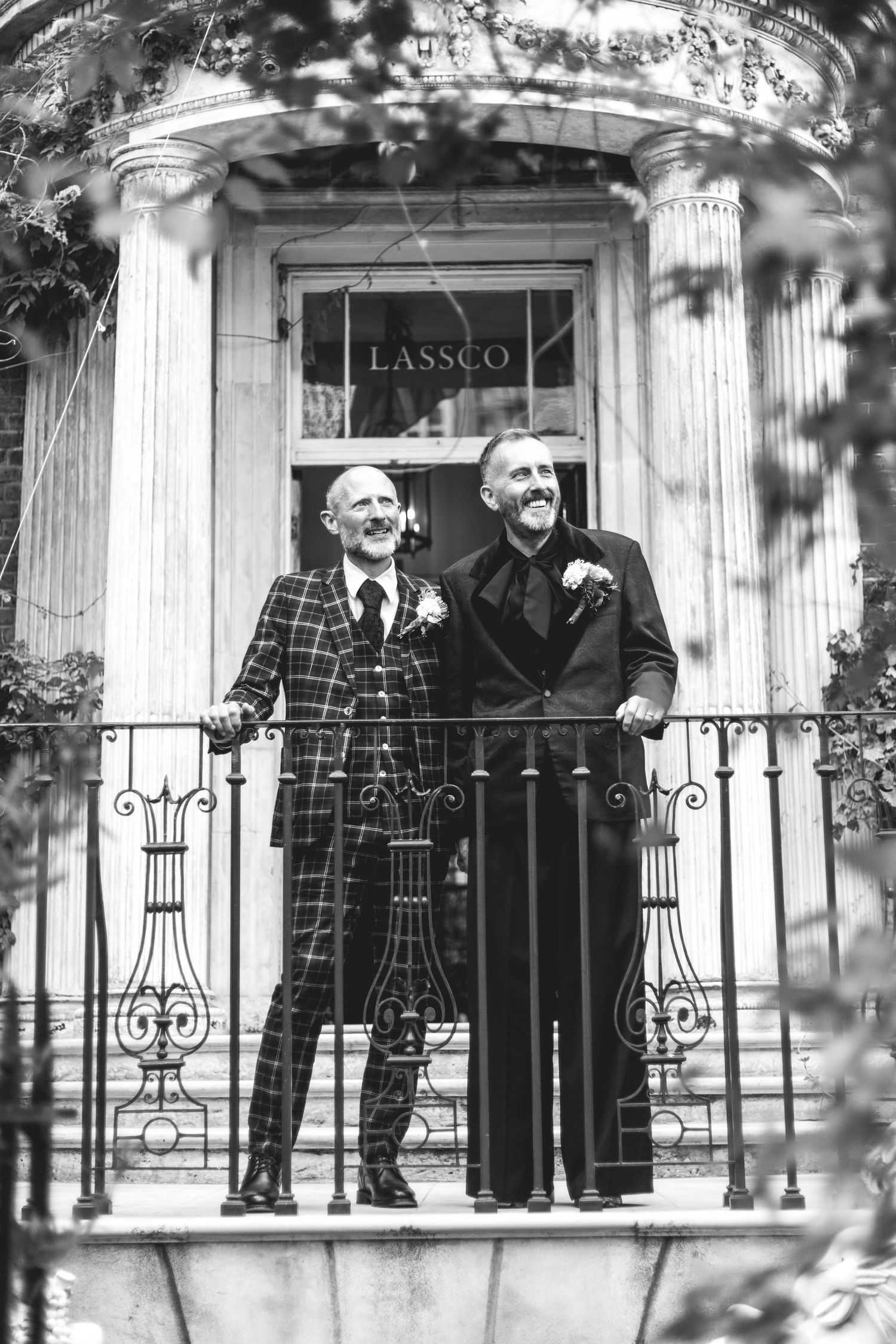 Two grooms in formal attire stand on a balcony outside LASSCO venue in this elegant black and white wedding photo.