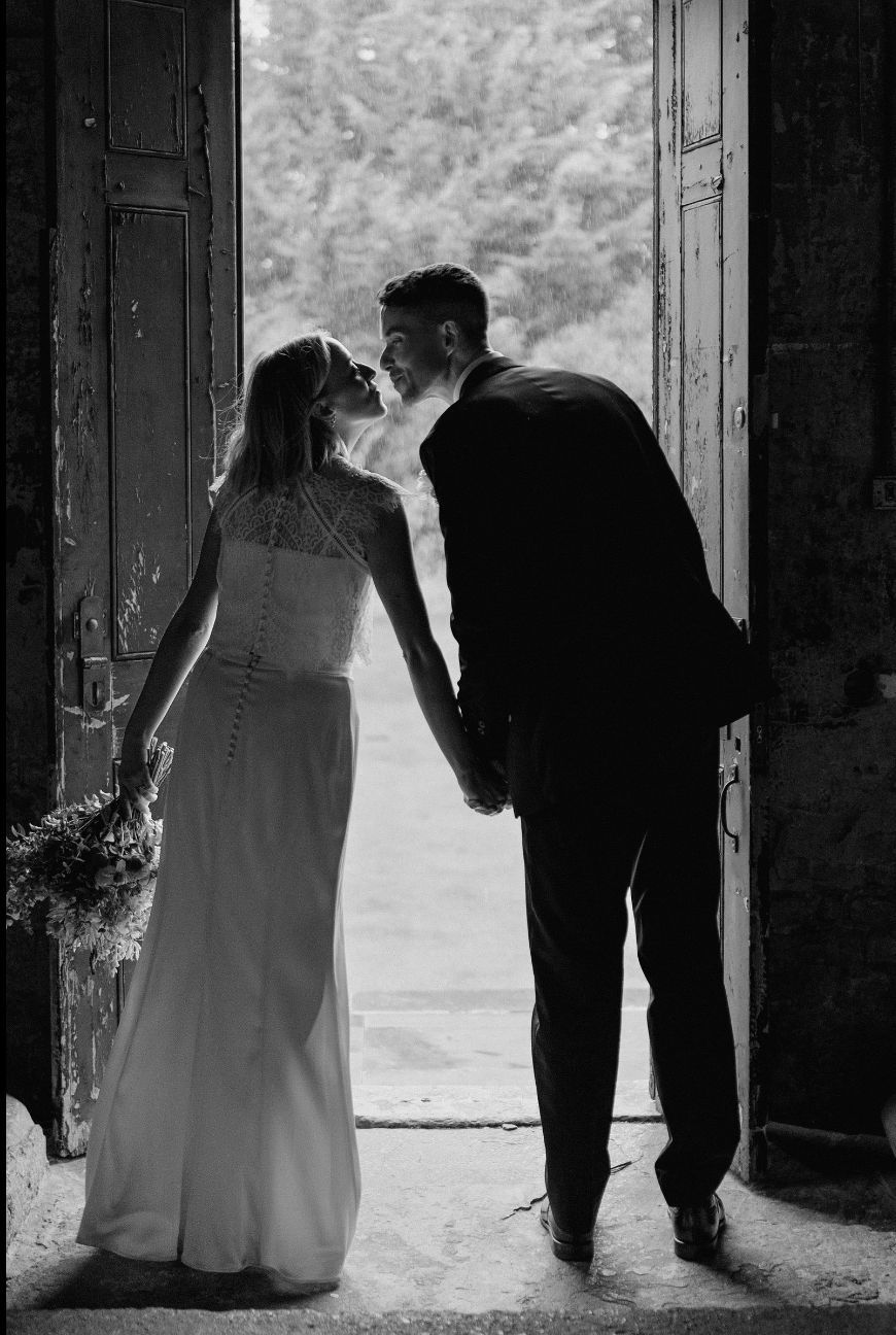 Black and white photo of a bride and groom holding hands, sharing a kiss in a rustic doorway on their wedding day.
