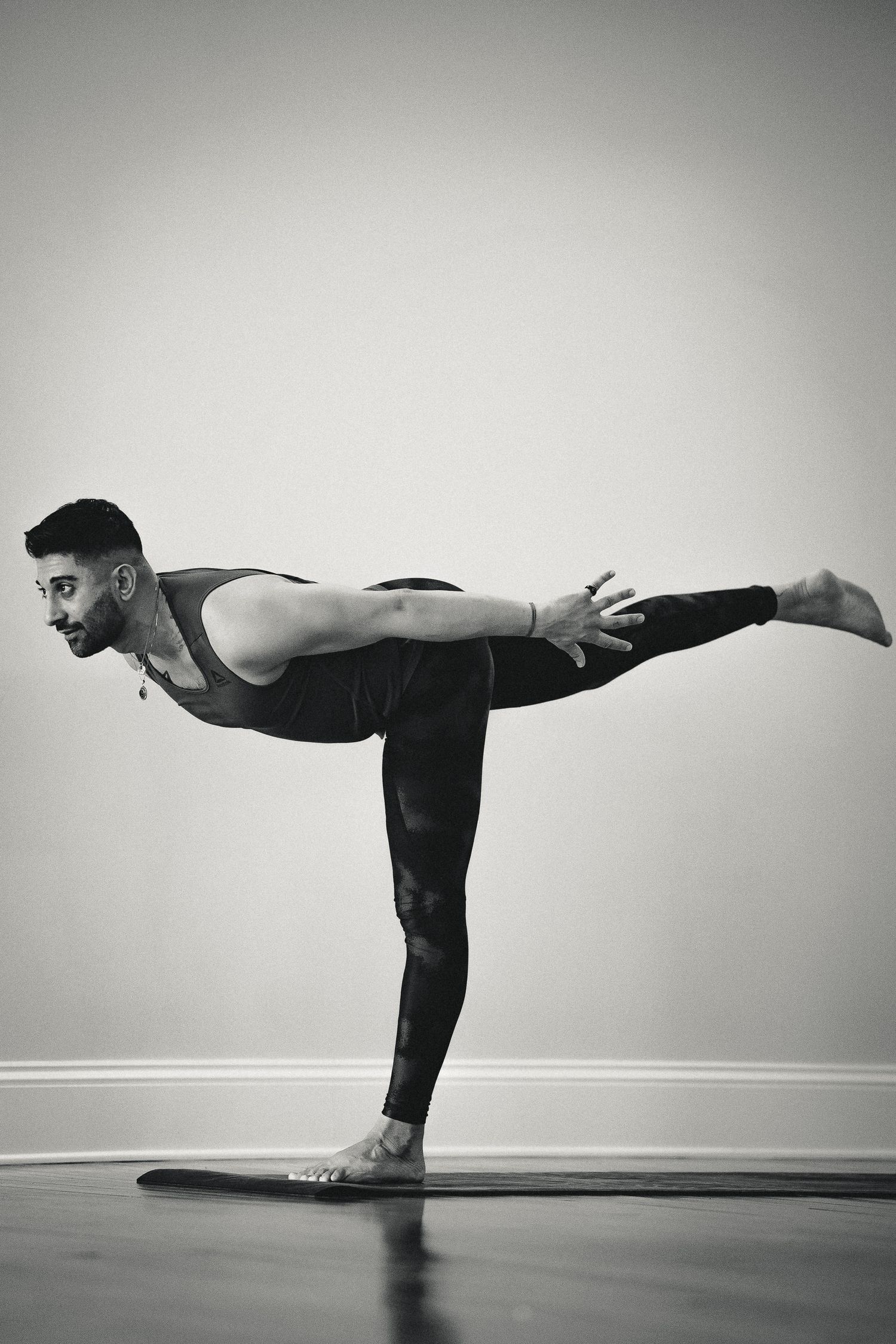 Male yogi performing Warrior III balance pose in black athletic wear on a mat against a neutral wall, black and white.