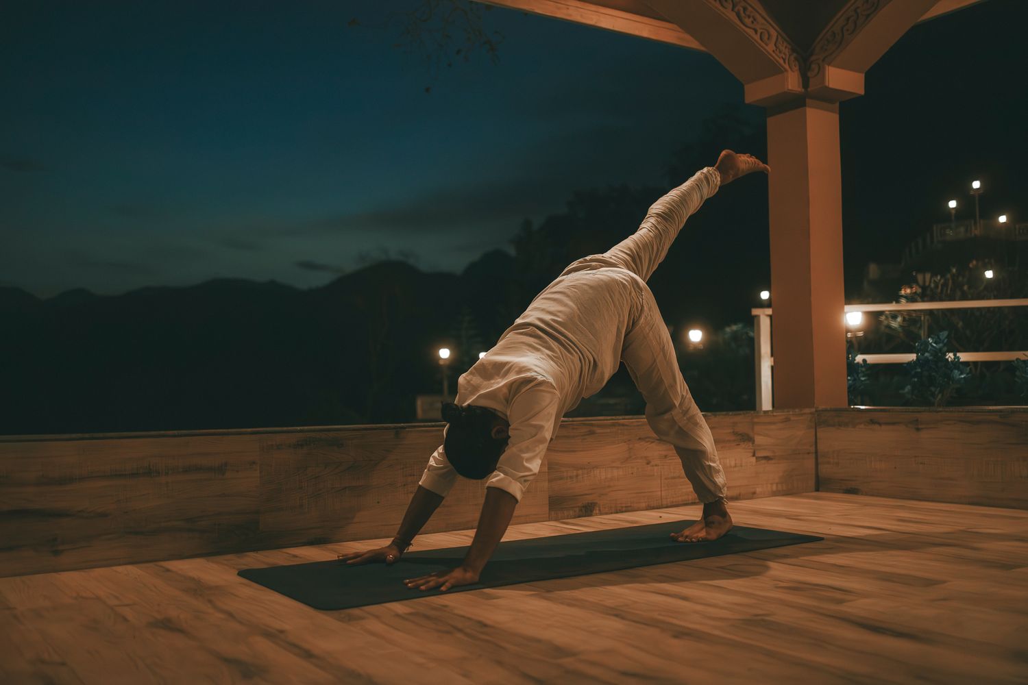 Person practicing wild thing yoga pose on a mat outdoors at night under a covered pavilion with city lights.