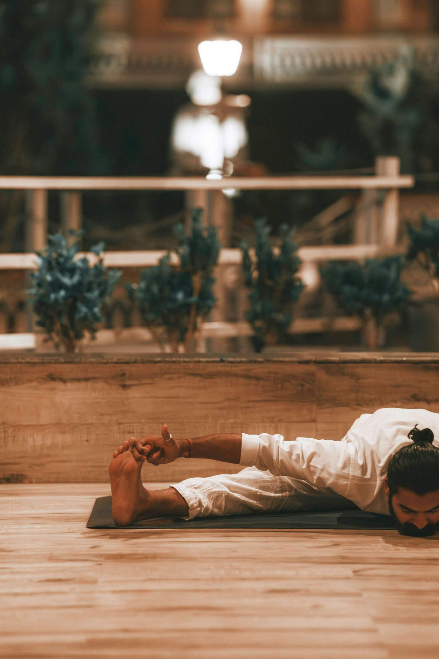 Man in white clothing performing a yoga bow pose on a mat on a wooden floor indoors.