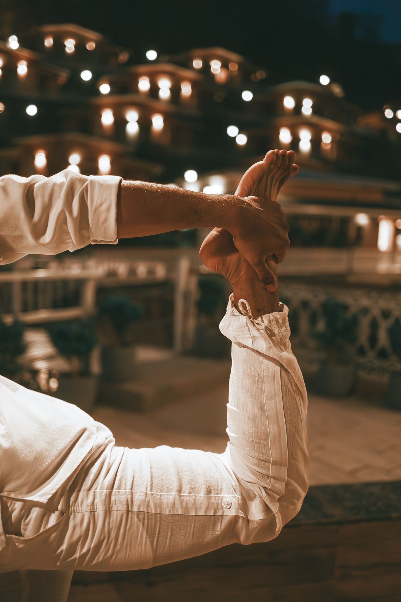 Close-up of a man in white performing a yoga bow pose holding his feet at night outdoors.