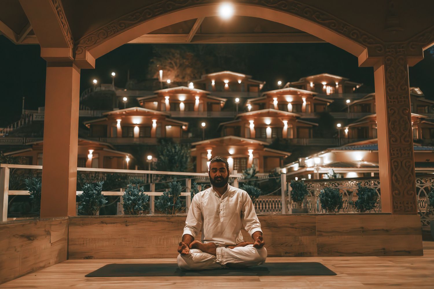 Bearded man in white meditating in lotus pose on yoga mat outdoors at night with lights.