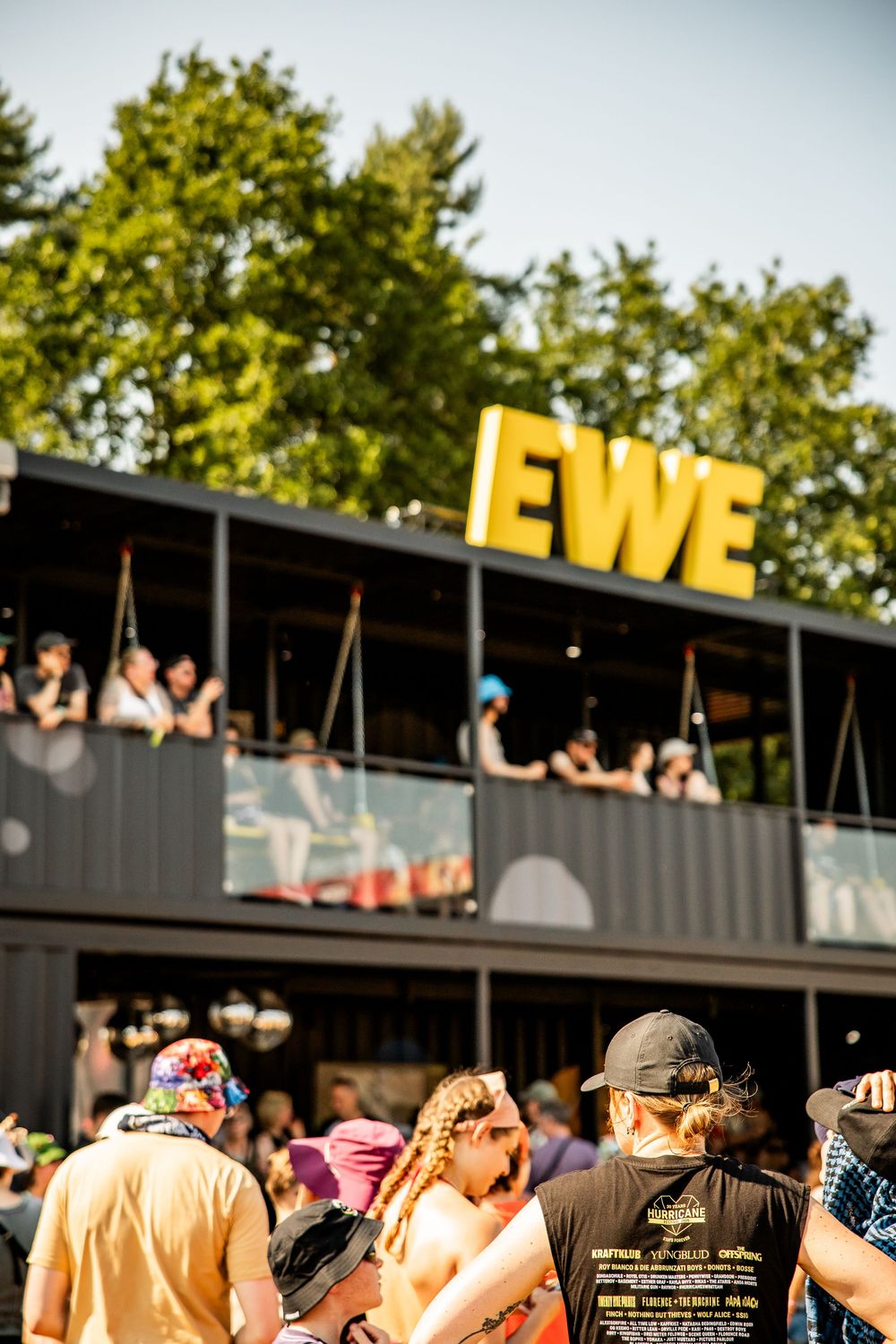 Crowd gathered at outdoor summer festival beneath bold yellow EWE sign on dark building.