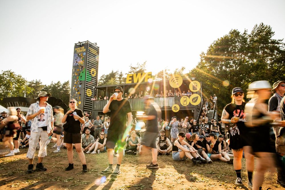 People dancing and sitting near the illuminated EWE Power Tower at a summer music festival at sunset.