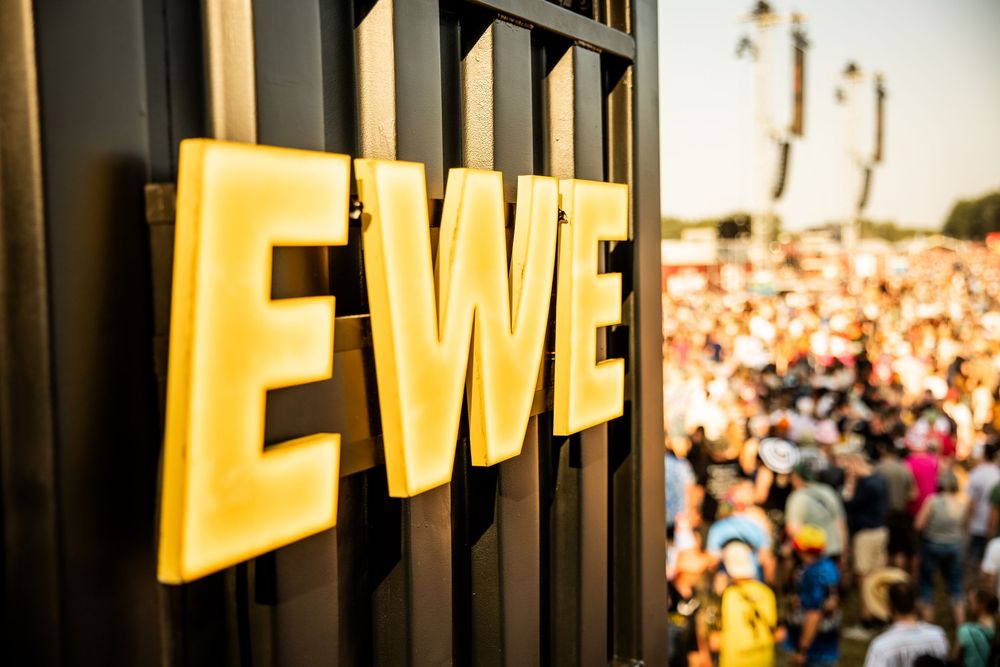 Close-up of illuminated yellow EWE letters sign with blurred festival crowd in the background.