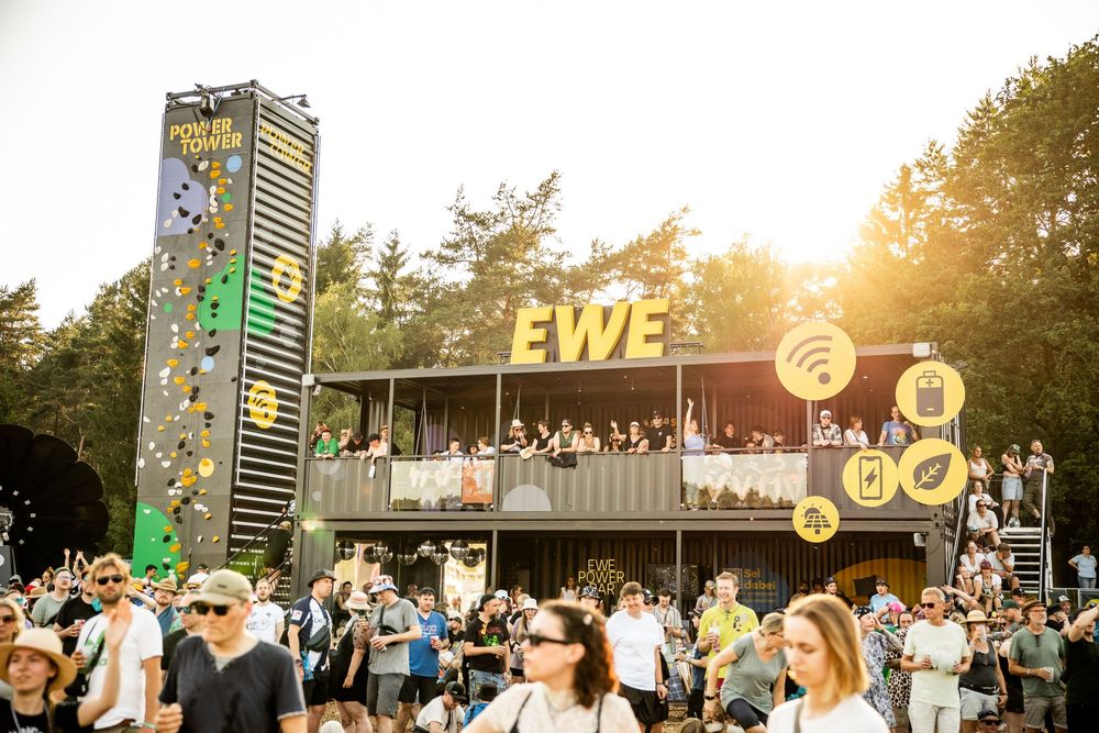 Crowd gathered at outdoor EWE festival stage with shipping container structure and climbing wall at sunset.
