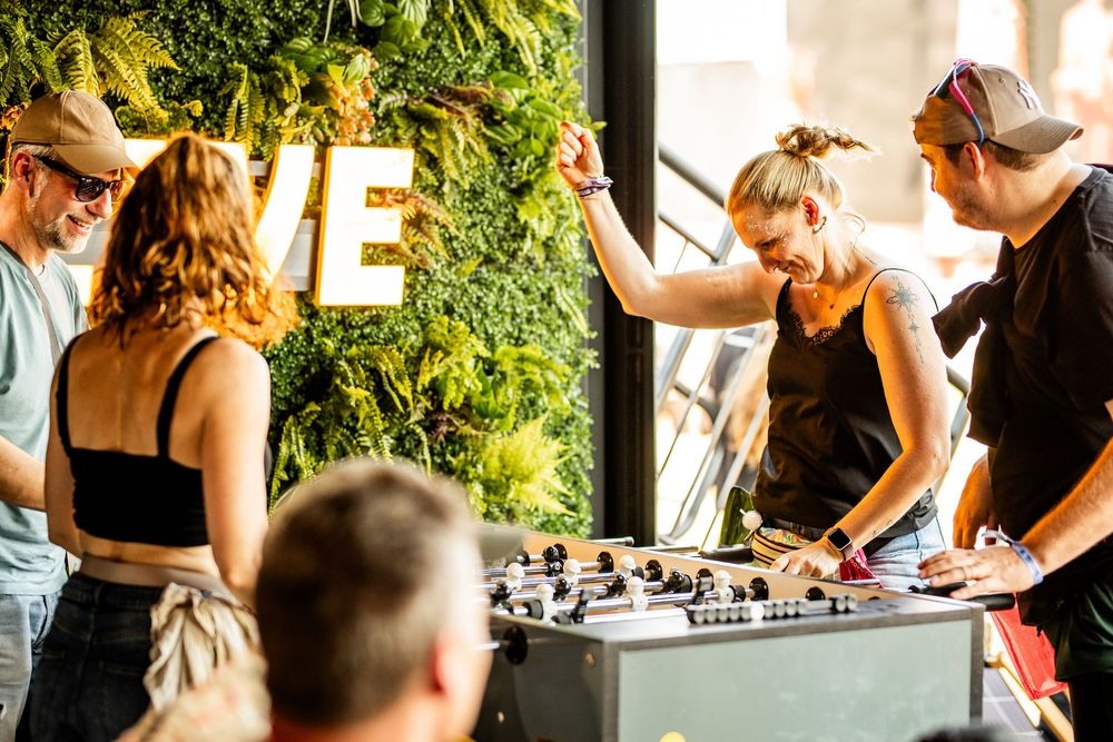 Woman playing foosball with friends indoors near green plant wall with illuminated VE sign.