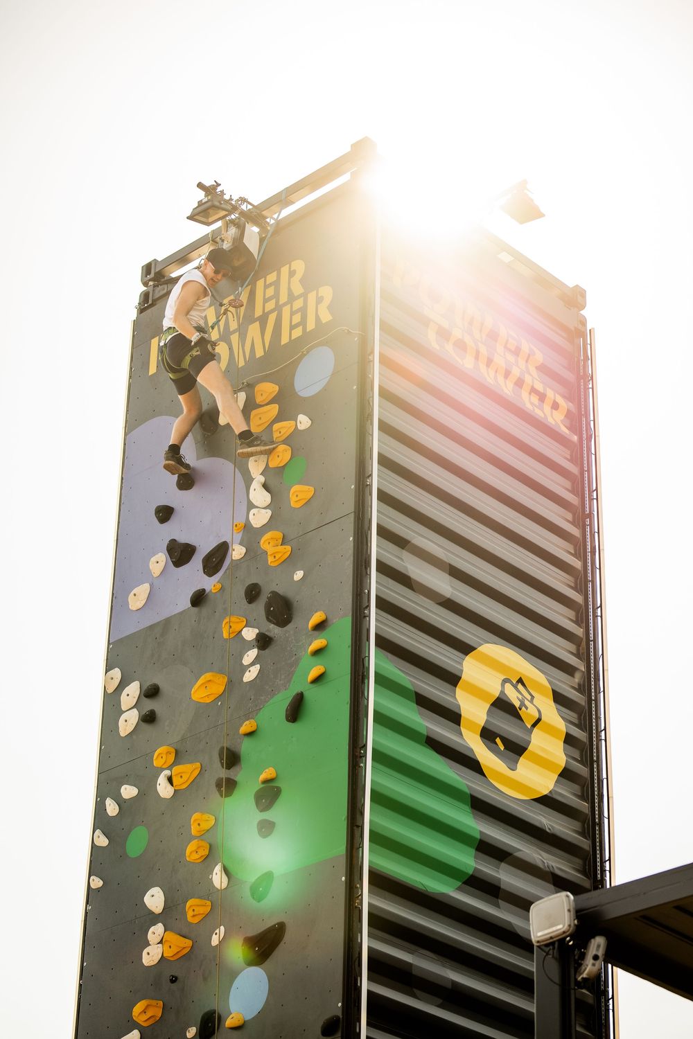 Athlete climbing tall outdoor rock climbing wall with bright sun flare at summer festival.