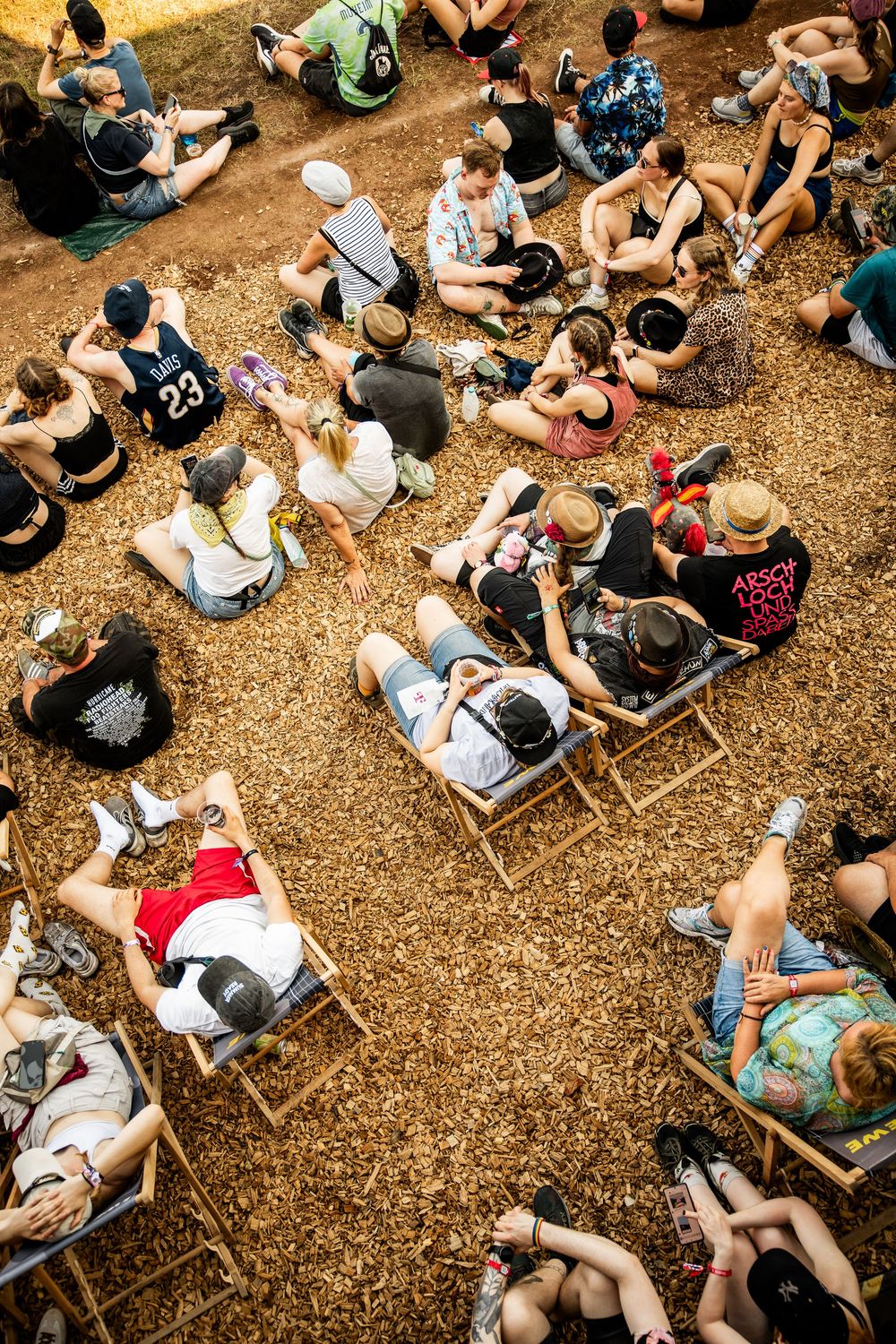 Aerial view of festival crowd sitting on wood chip ground in deck chairs at a summer music event.