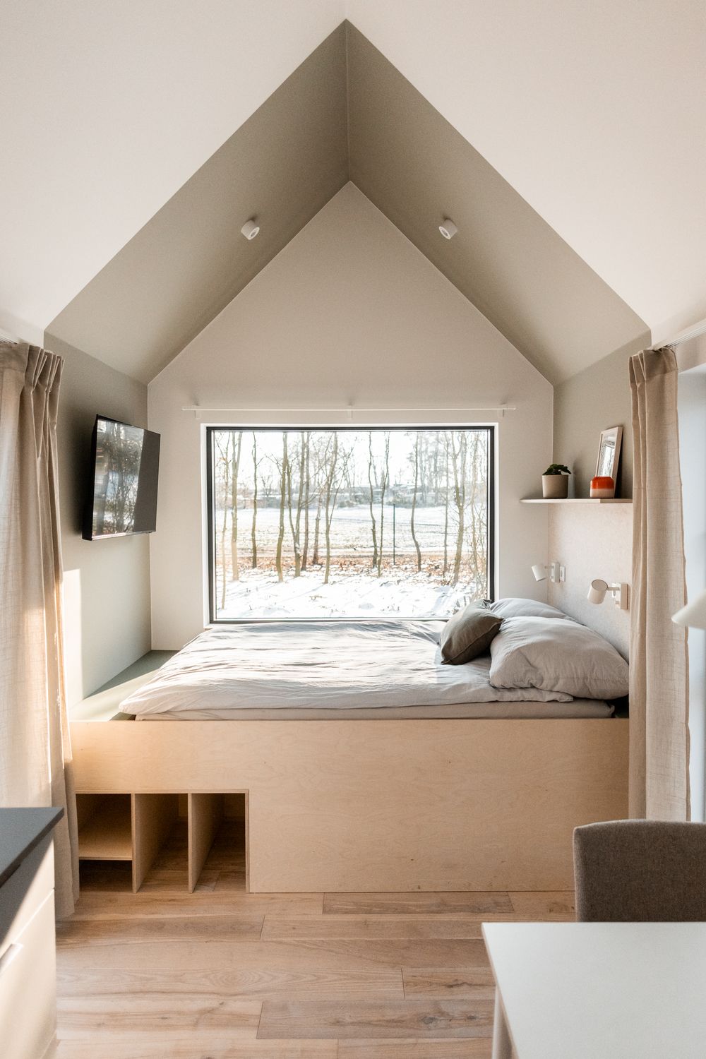 Minimalist tiny house bedroom with platform bed, built-in storage, vaulted ceiling, and large window overlooking snowy trees.