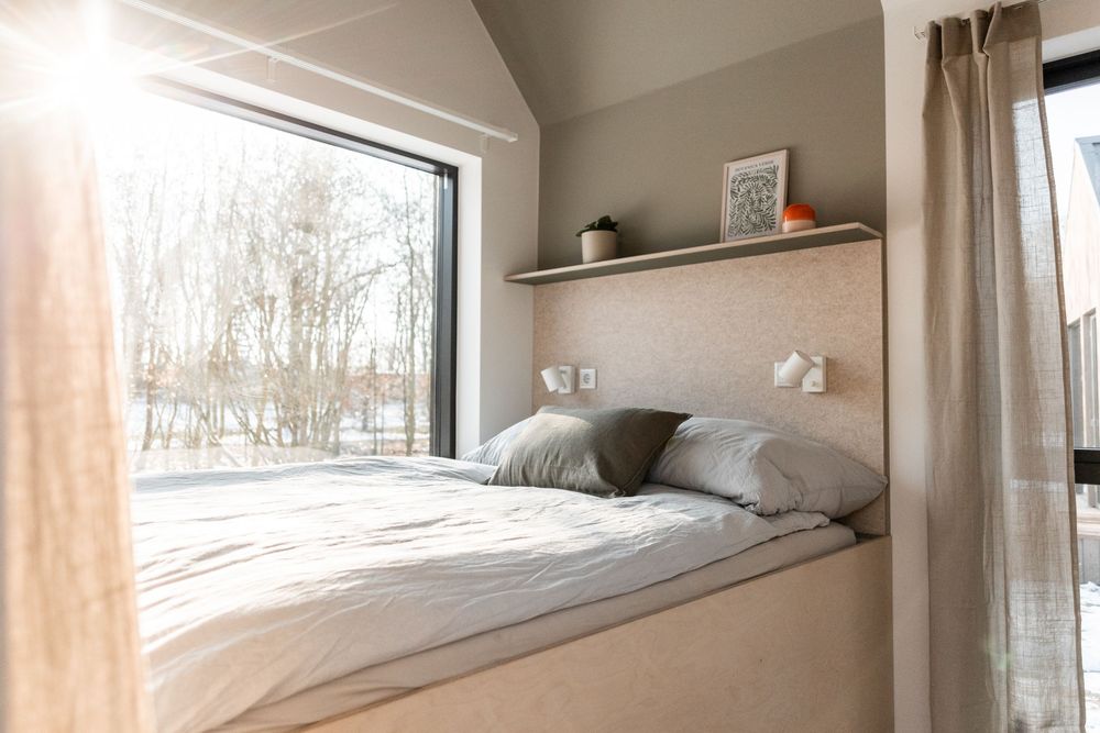 Cozy modern bedroom with neutral bedding, wall shelf, reading lights, and large window with winter forest view.