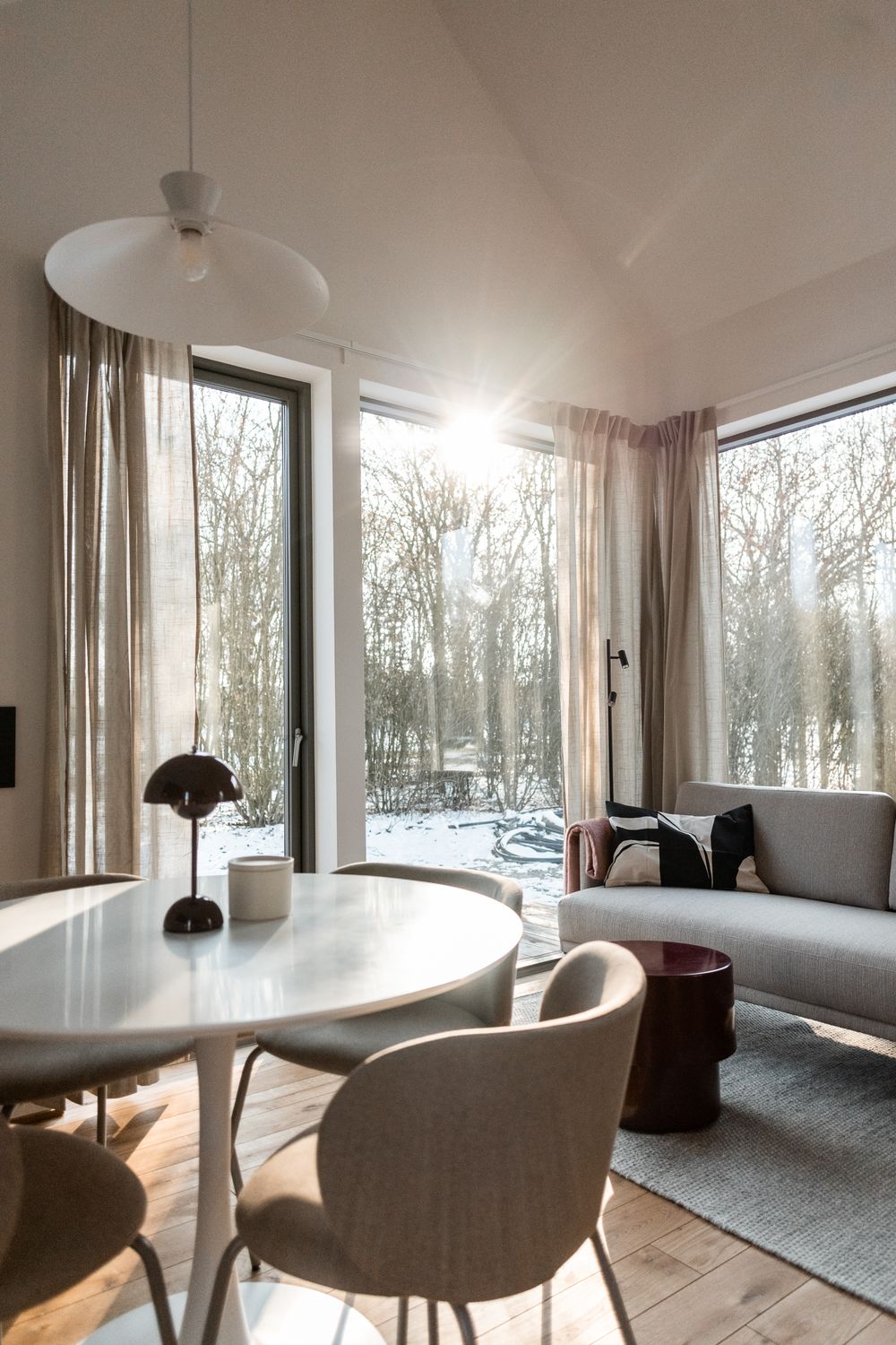 Bright Scandinavian living area with round dining table, gray sofa, large windows, and sunlight streaming through winter trees.