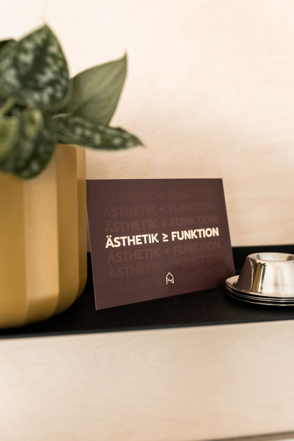 Dark business card reading Asthetik gleich Funktion displayed on black shelf beside gold planter and small bowl.