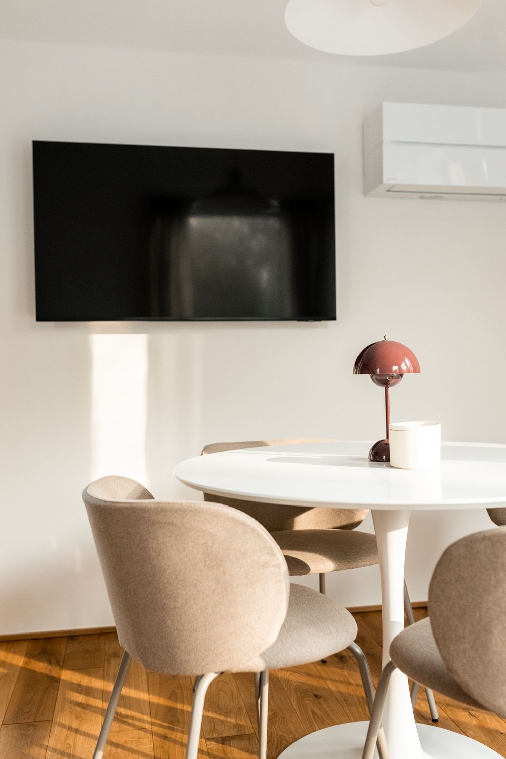 Modern dining area with round white table, beige chairs, wall-mounted TV, and red Flowerpot lamp on wood floor.