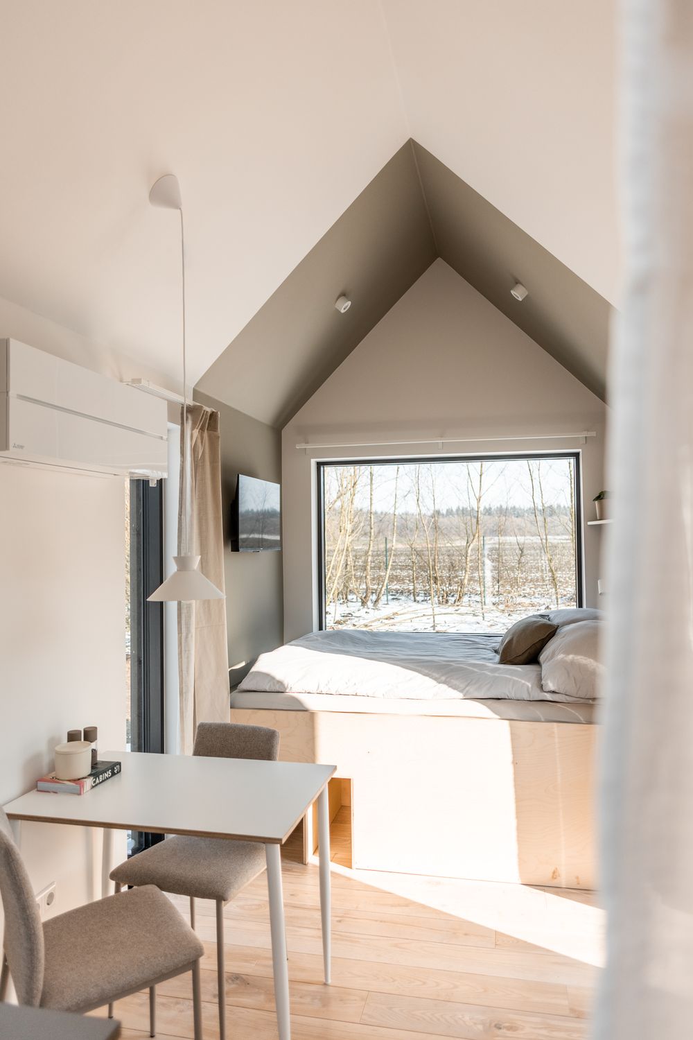 Bright minimalist cabin interior with peaked ceiling, built-in bed platform, and panoramic nature window.