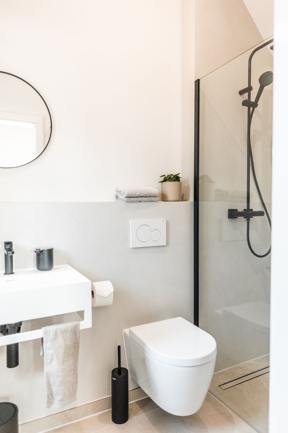 Modern compact bathroom with wall-mounted toilet, glass shower, round mirror, and black fixtures.