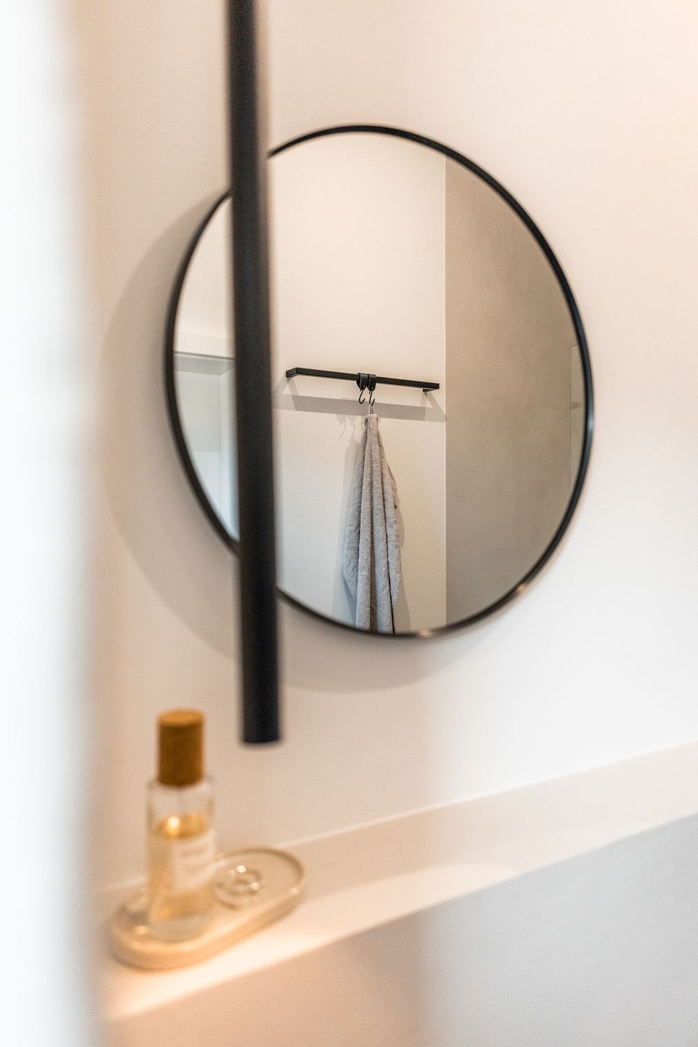Round black-framed mirror reflecting coat hook in minimalist white bathroom with perfume bottle on shelf.