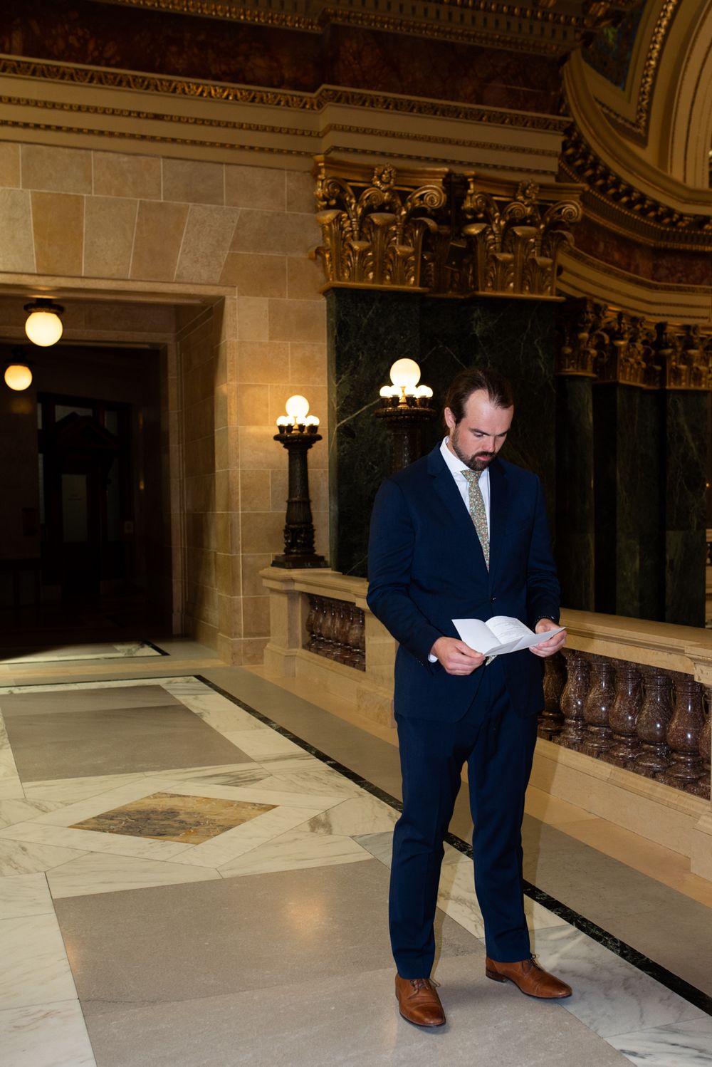 Groom in navy suit reading vows in ornate marble hall.