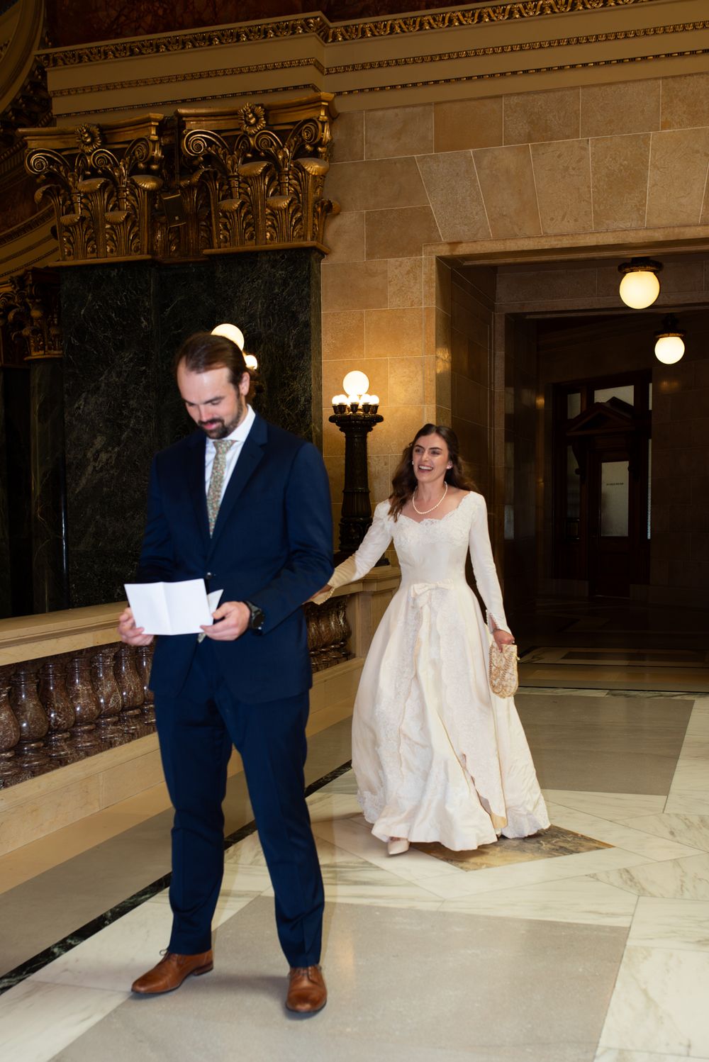 Bride in ivory gown reaches for groom in navy suit walking through ornate marble hall.