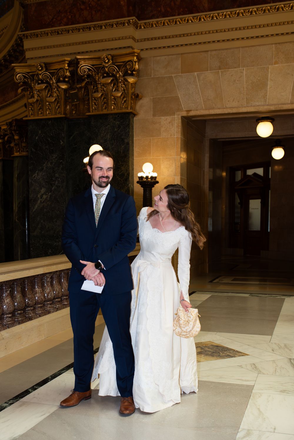 Bride in white long-sleeve gown and groom in navy suit walk together smiling in ornate marble building.