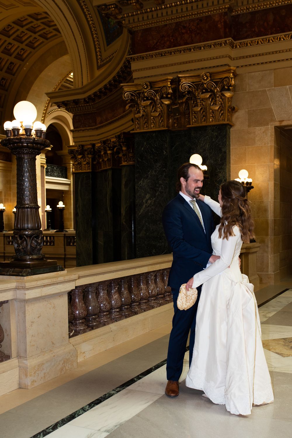 Bride and groom share tender moment in ornate wedding venue with grand marble columns.
