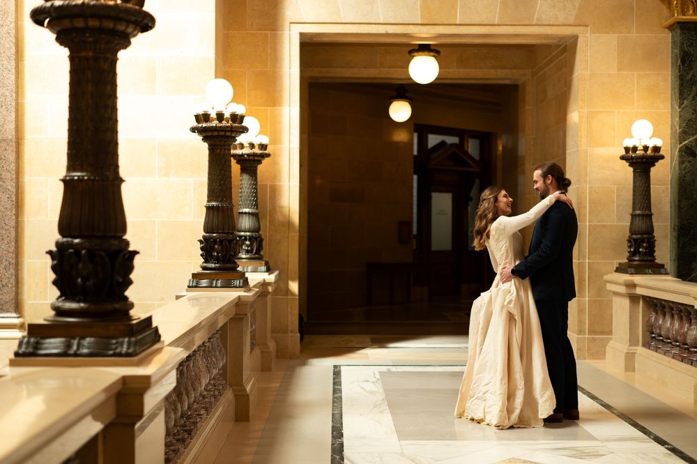 Couple embraces romantically in elegant golden-lit hall with decorative pillars on wedding day.