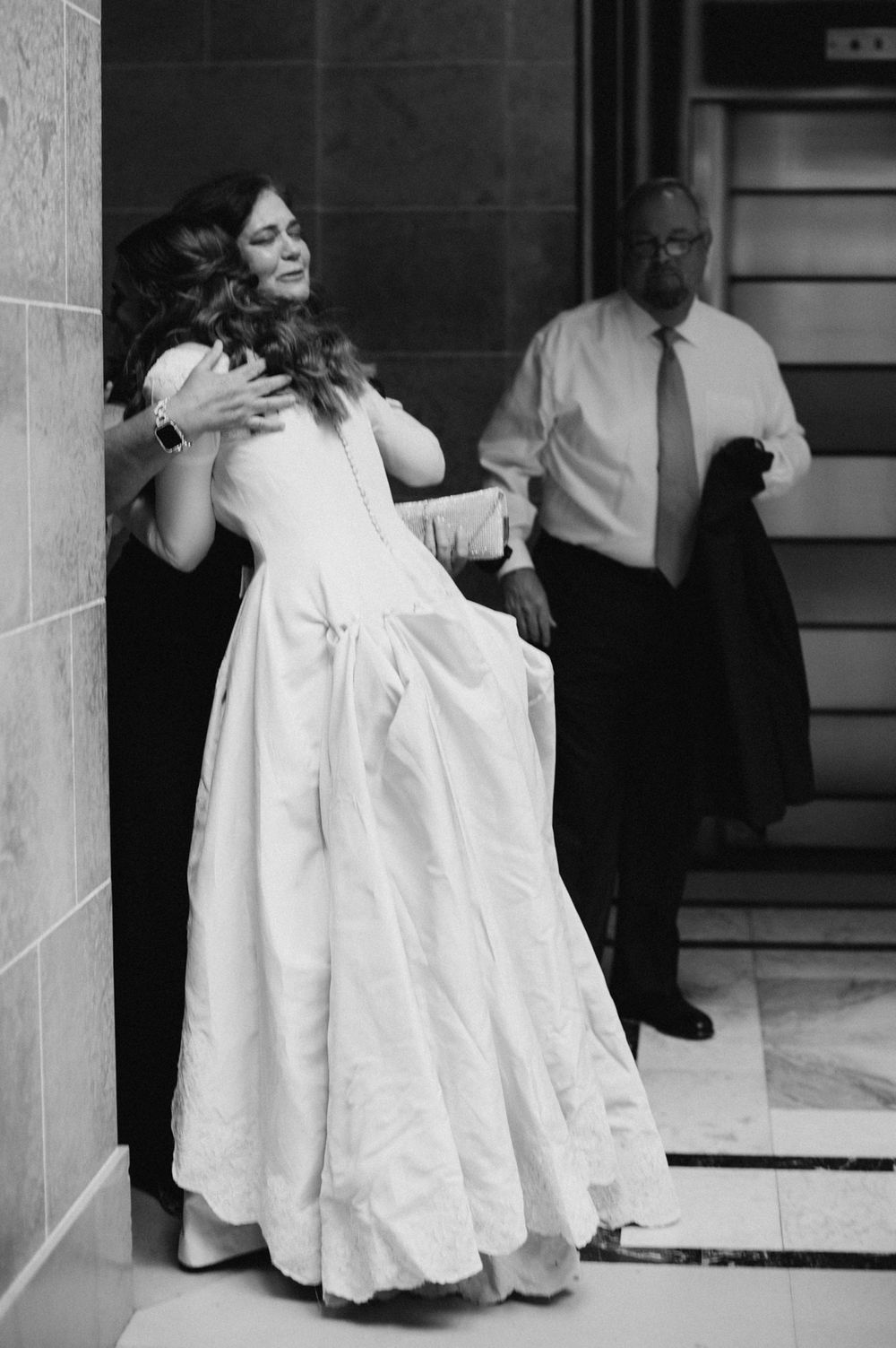 Bride in white gown receives hug while man in tie watches in black and white wedding photo.