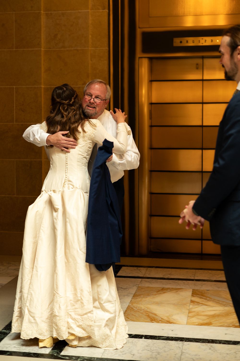 Bride in ivory gown embracing older man in navy suit near elevator in elegant marble venue.